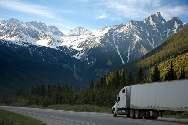 Bílý nákladní automobil jede po cestě mezi horami a přírodou, kolem zasněžených vrcholků hor.